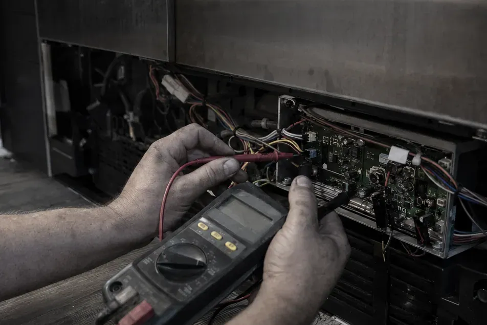 Hands holding a multimeter and test leads at the control board of an open refrigerator base in a dim room.