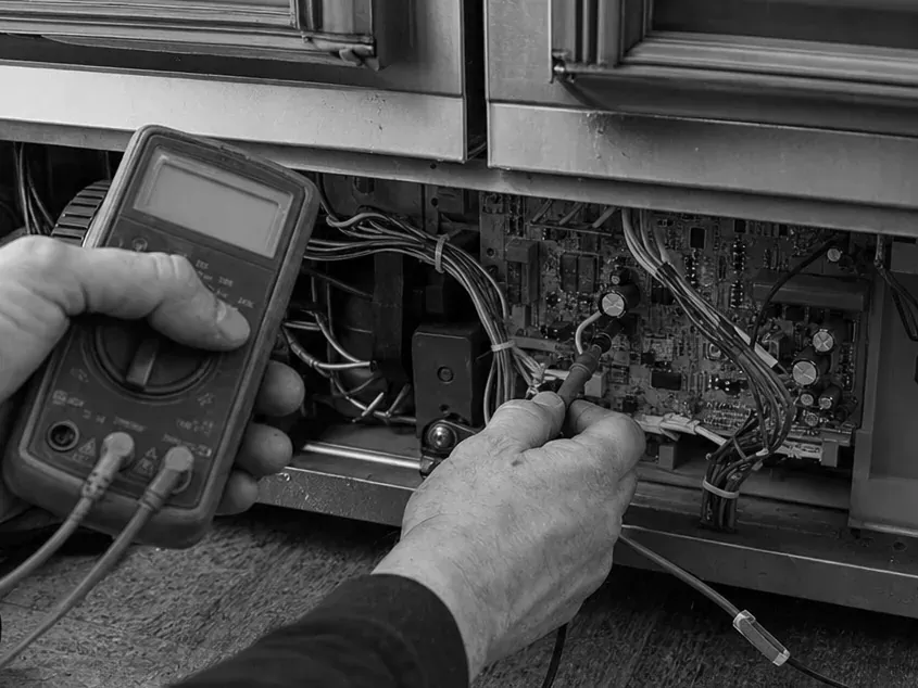Black-and-white view of hands holding a multimeter and probing the control board at the base of a glass-door refrigerator.