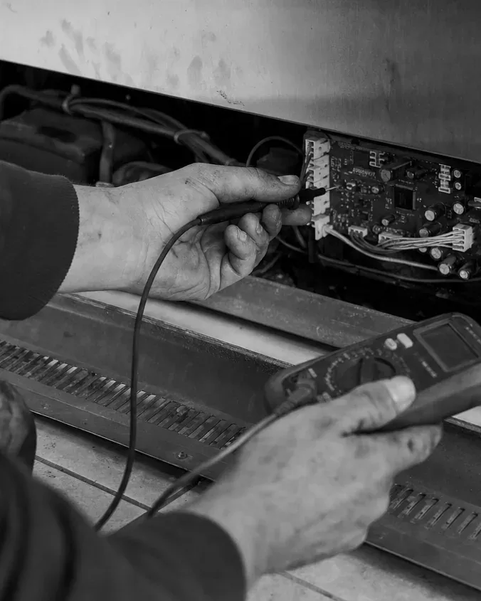 Black-and-white view of a technician's hands with a multimeter at the control board behind a refrigerator's kick grille.